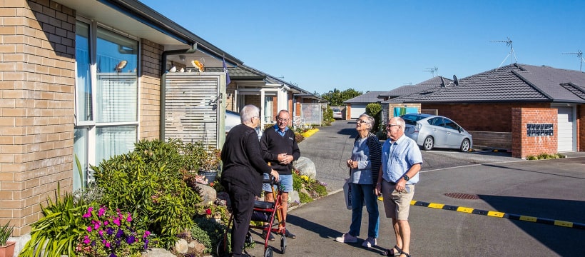 Residents engaging in conversation outside private apartments at a senior living community, featuring accessible pathways and landscaped gardens, highlighting social interaction and community living.
