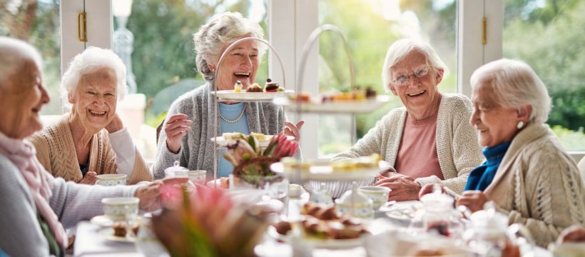 Group of elderly women enjoying a social gathering over tea and pastries in a bright, inviting setting, reflecting the community and socialization aspects of independent living at Dorset Place Senior Living.